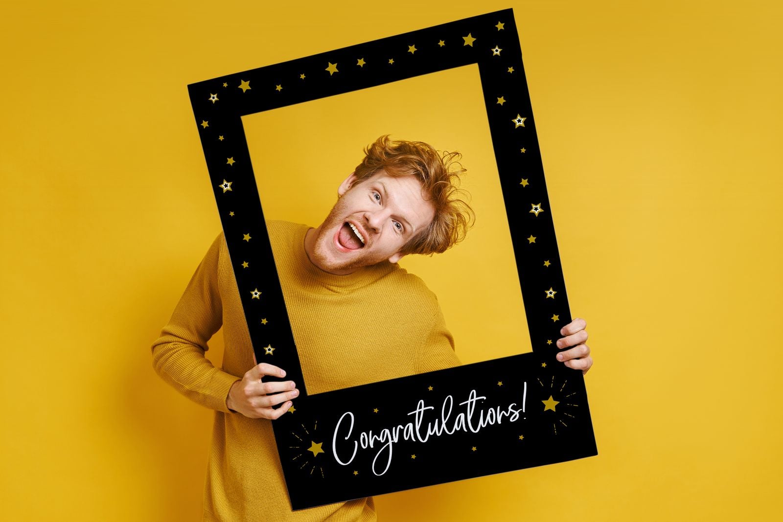 Man posing in a black “Congratulations!” selfie frame against a yellow wall